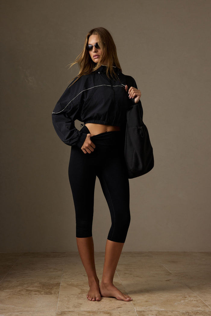 Model wearing black Acadia cropped leggings and cropped windbreaker, holding black bag and adjusting waistband in studio