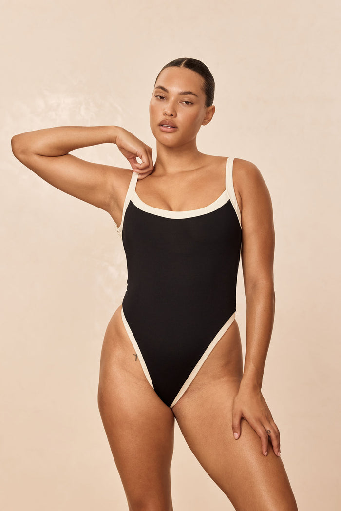 Front view of Barcelona One Piece in black with ivory trim on model, high-cut leg and scoop neckline against beige background