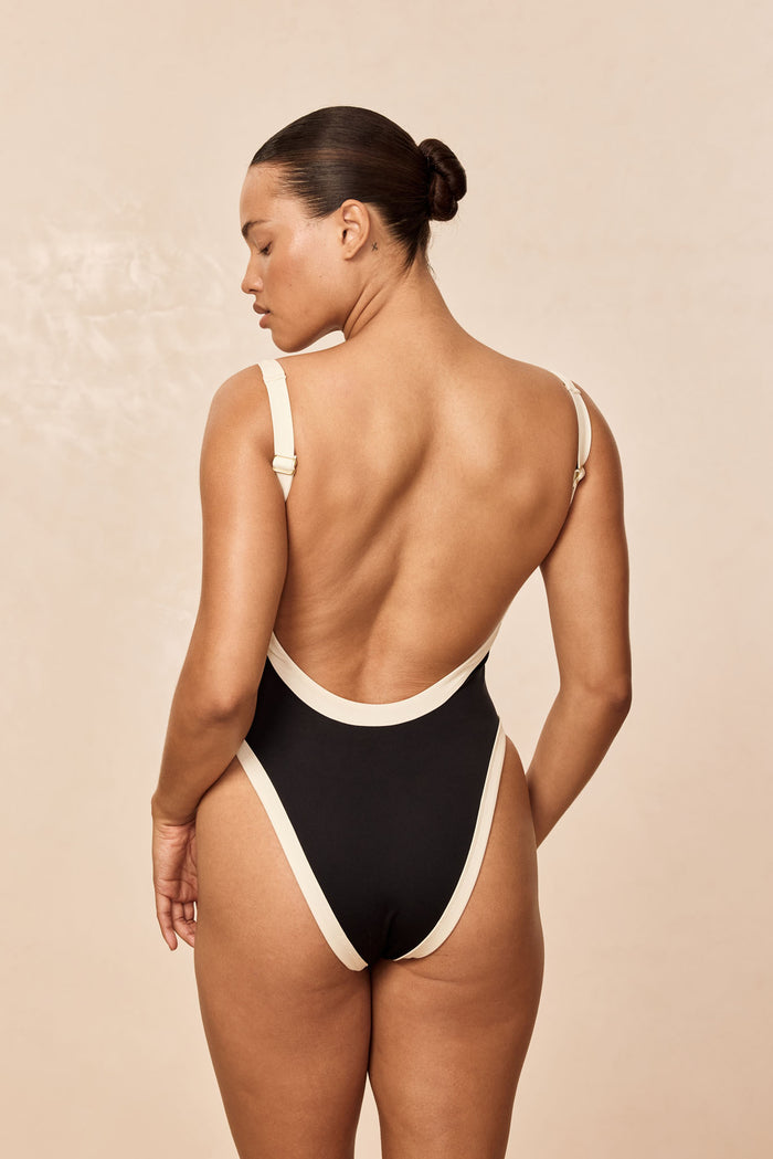 Back view of Barcelona One Piece in black and ivory showing low scoop, high-cut leg and adjustable straps on beige