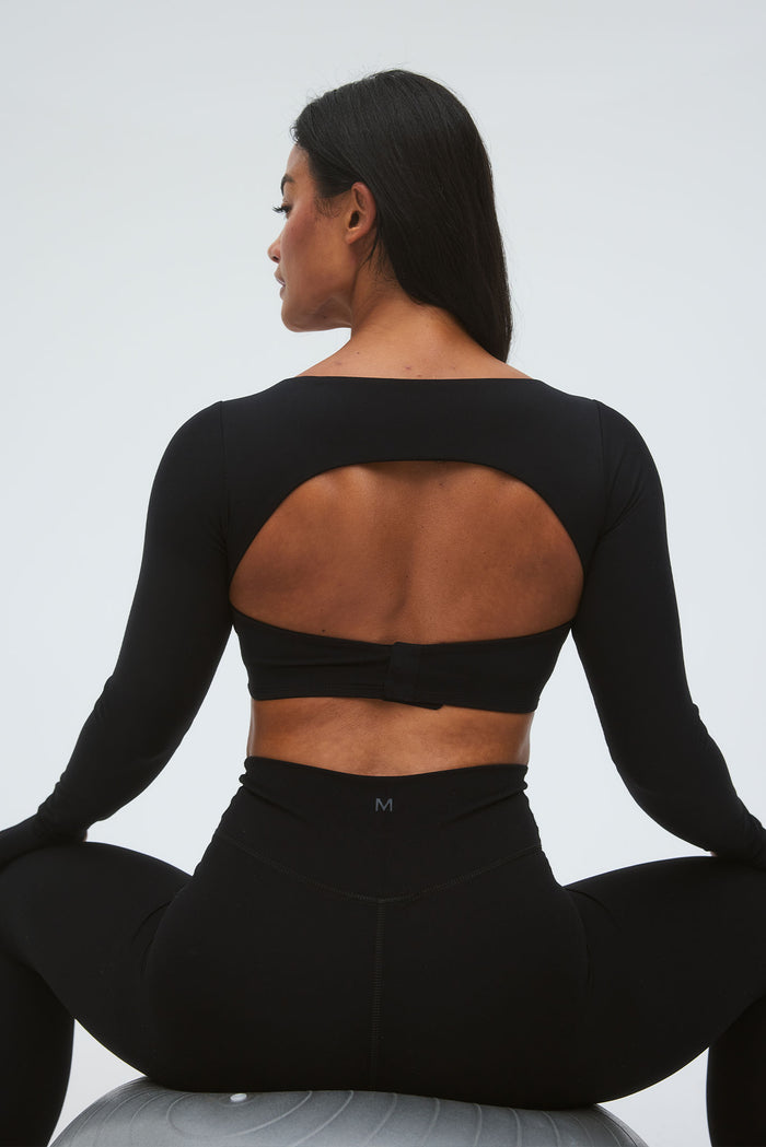 Back view of model wearing Bellevue Top in black, fitted long-sleeve crop top with oval back cutout and adjustable hook-and-eye band, seated on a gray exercise ball