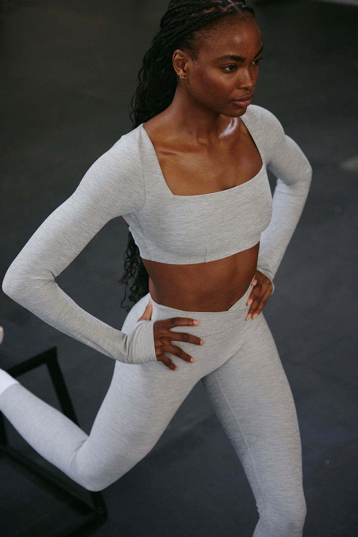 Three-quarter view of Bellevue Top in Heather Grey showing square neckline, long sleeves with thumb holes, and cropped fit on a model against a dark studio floor
