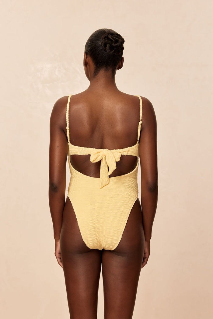 Back view of Clovelly One Piece in honey crinkle showing tie-back cutout, adjustable straps, high-cut leg on beige background