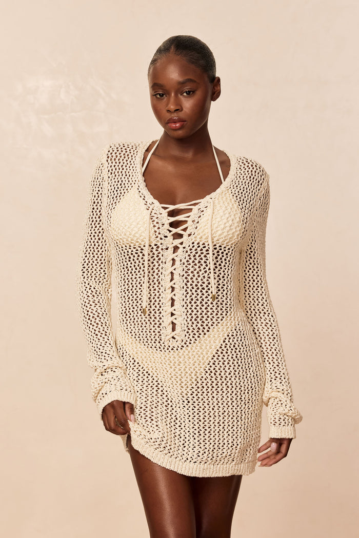 Front view of Corossol mini dress in ivory petit crochet worn over bikini, lace-up neckline, open-knit long sleeves and mini length with neutral beige background