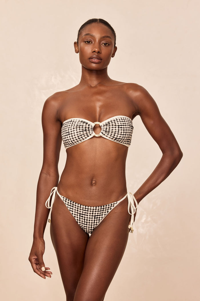 Model wearing strapless gingham bikini with gold ring center and tie-side bottoms, neutral studio background.