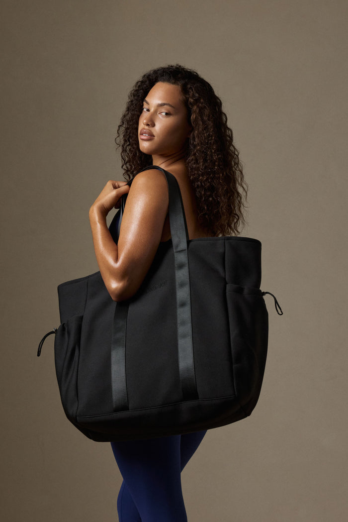 Model wearing black Monday Body tote over shoulder, oversized with exterior drawcord pockets and tonal MONDAY logo