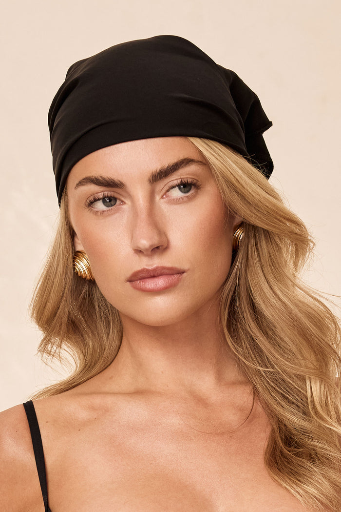 Front view of Monday Headscarf in black on model, smooth rayon square scarf tied at the back against a neutral beige background