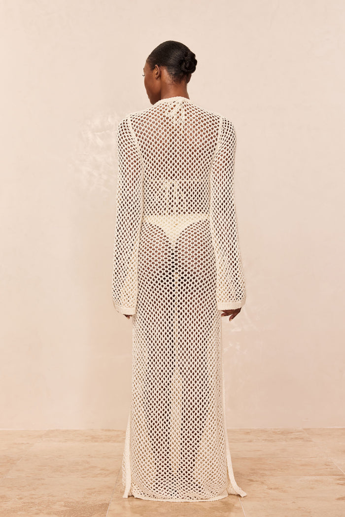 Back view of Montego Dress in ivory chunky crochet open-knit maxi with long sleeves over swimwear on neutral beige background