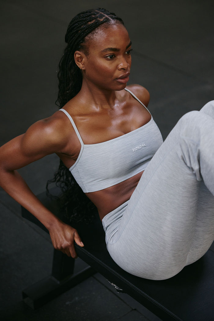 Front view of Olympic Bra in Heather Grey with scoop neckline, adjustable straps and MONDAY embroidery on model on a bench