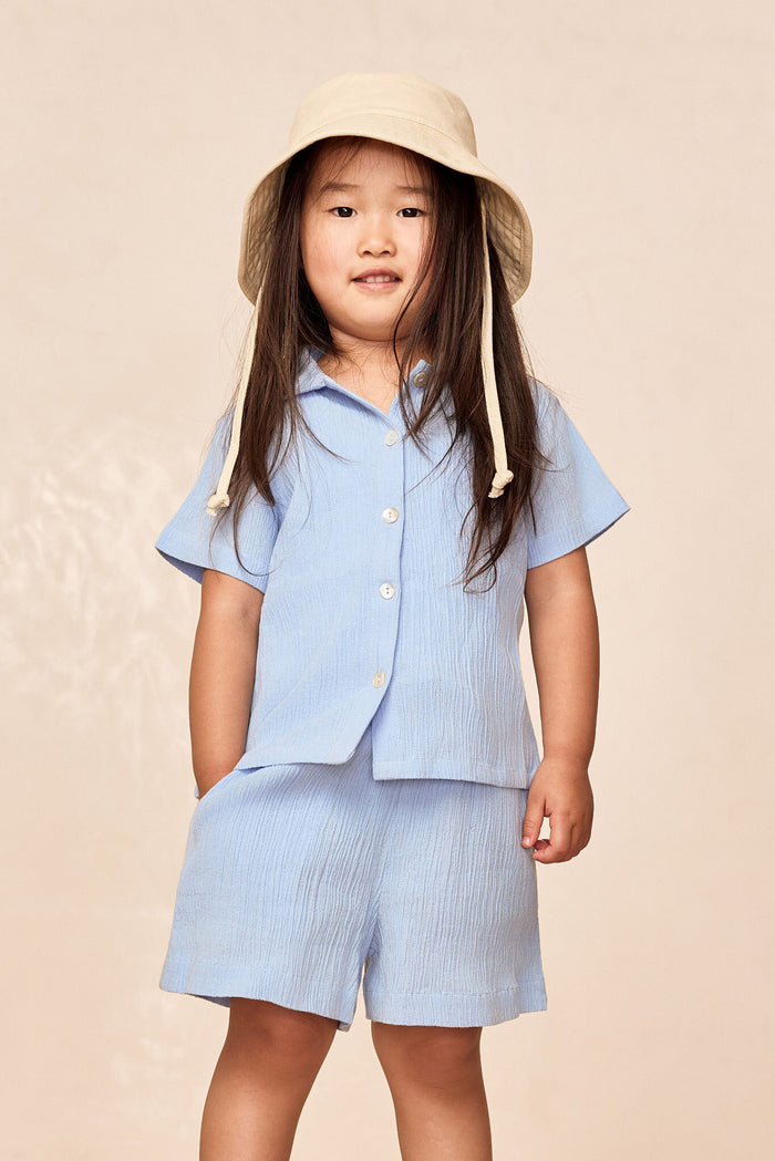 Front view of Pacific Beach Shirt Kids in Sky showing button-front, short sleeves and crinkled fabric on child model with beige background