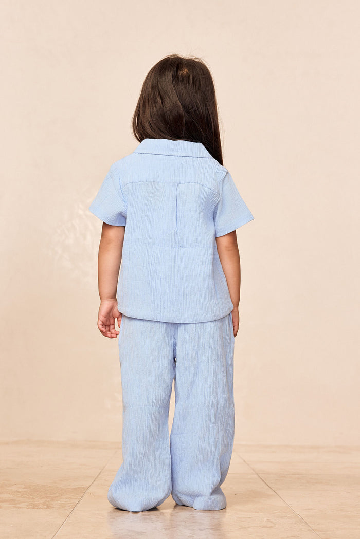 Back view of Pacific Beach Shirt Kids in sky blue showcasing crinkle rayon-linen fabric and short sleeves on a neutral beige studio background