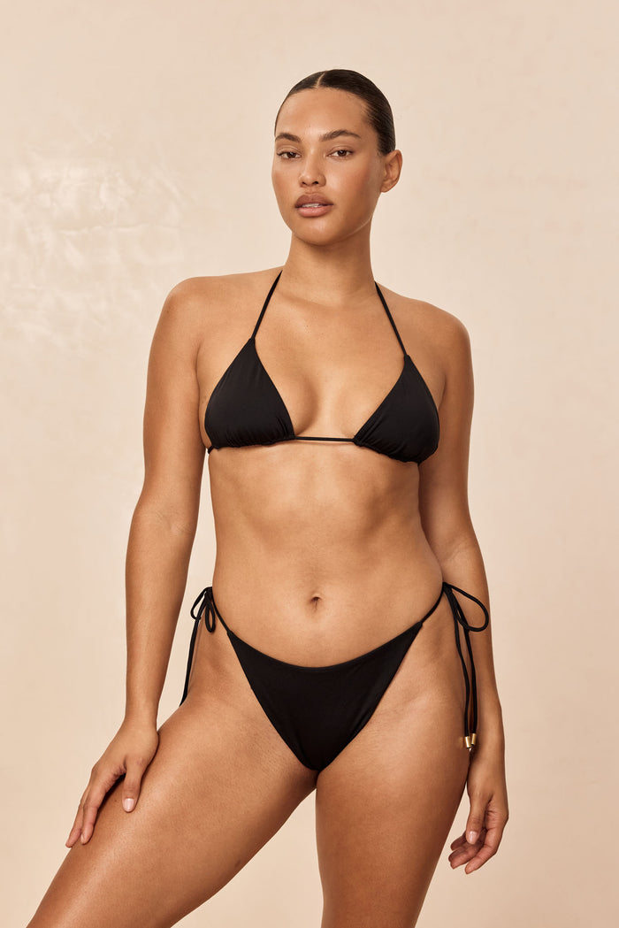 Front view of Taormina Top in black showcasing triangle halter shape and thin tie straps on model against beige background