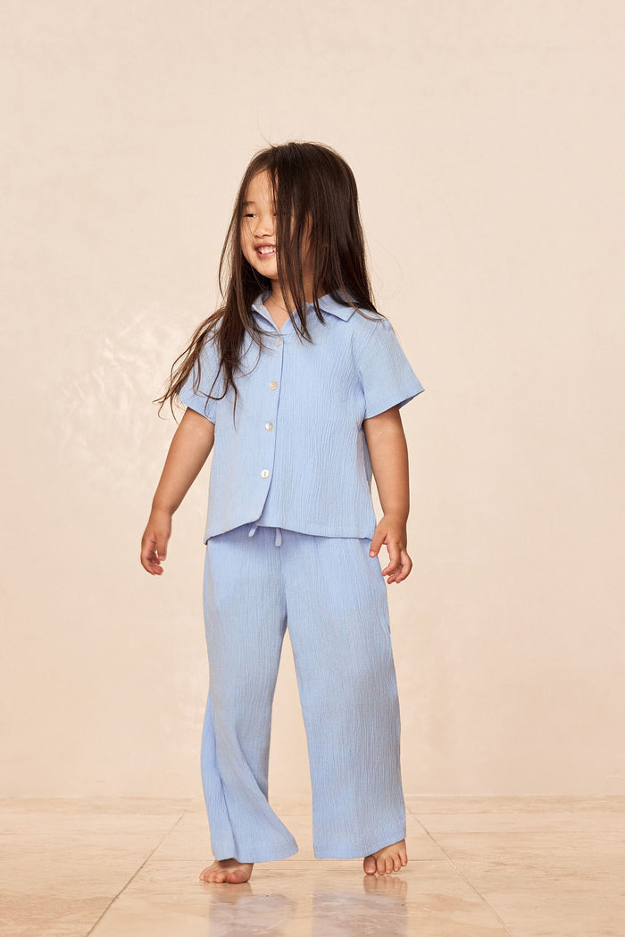 Three-quarter view of Tuscany Pants Kids in Sky on child, showing smocked waistband and flared legs on beige studio background